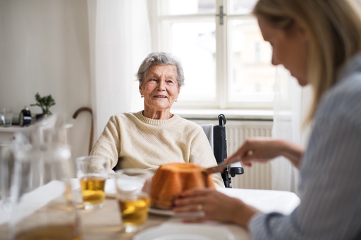 An elderly woman sat in a wheelchair at a table smiling while a carer cuts cake