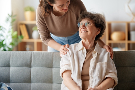 An old lady sat on a sofa with a carer behind her, placing her hands on her shoulders gently