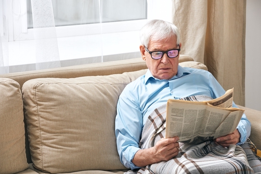 An elderly man with a blanket over his lap sat on a sofa reading a newspaper